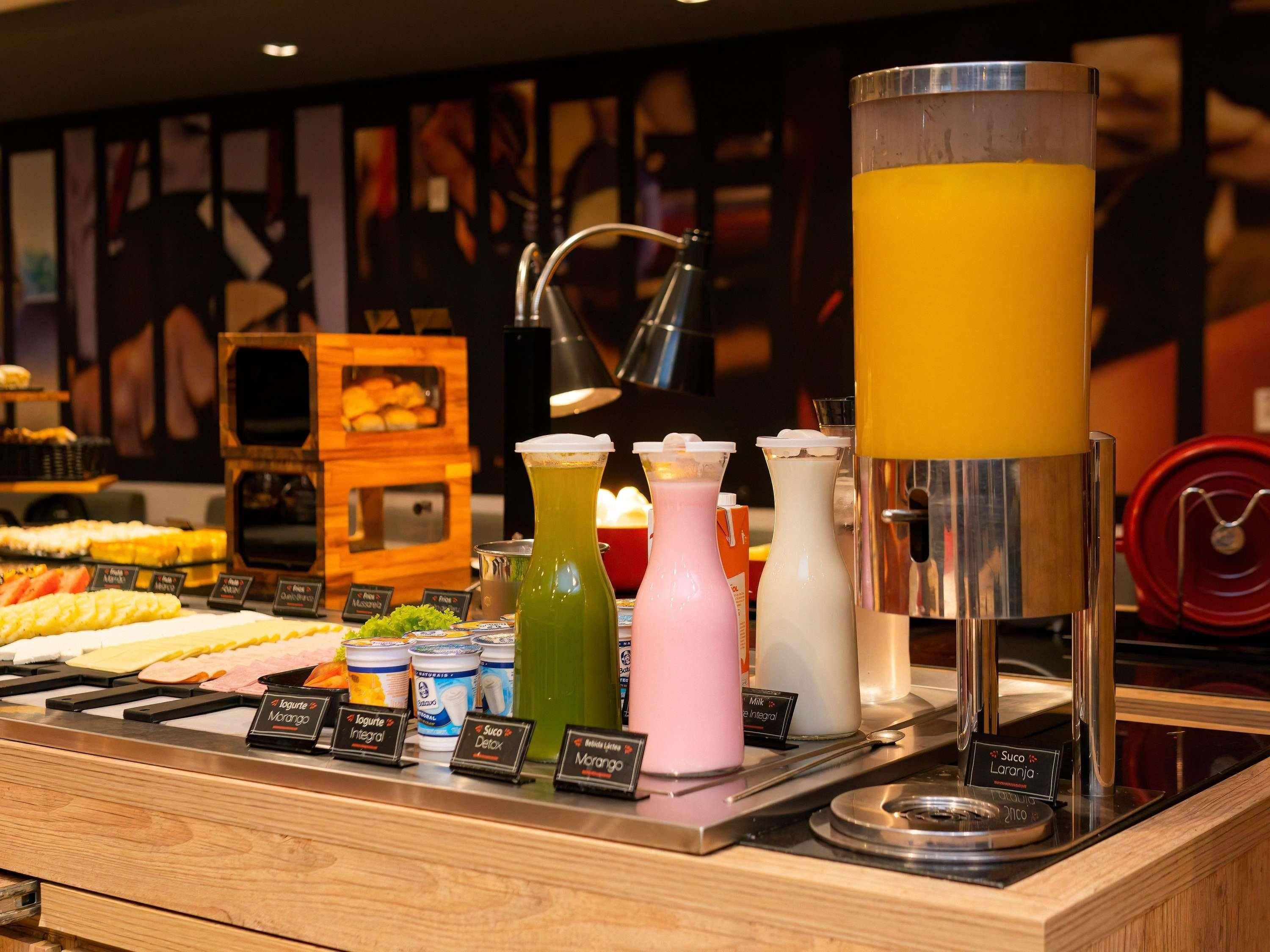 Buffet com diversos alimentos e bebidas, incluindo um grande dispensador amarelo para líquidos e vários recipientes rotulados de iogurte e leite.