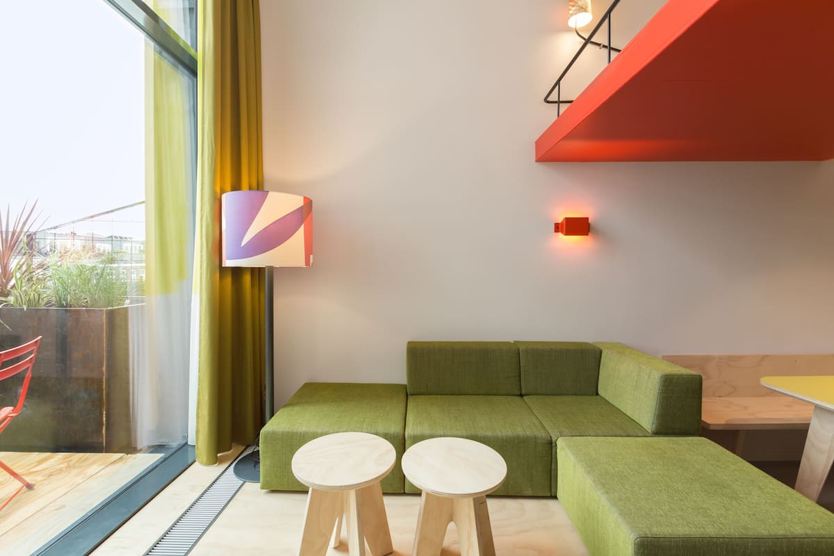 Una sala de estar moderna con un sofá seccional verde, dos taburetes de madera, una mesita y un amplio ventanal con cortinas.