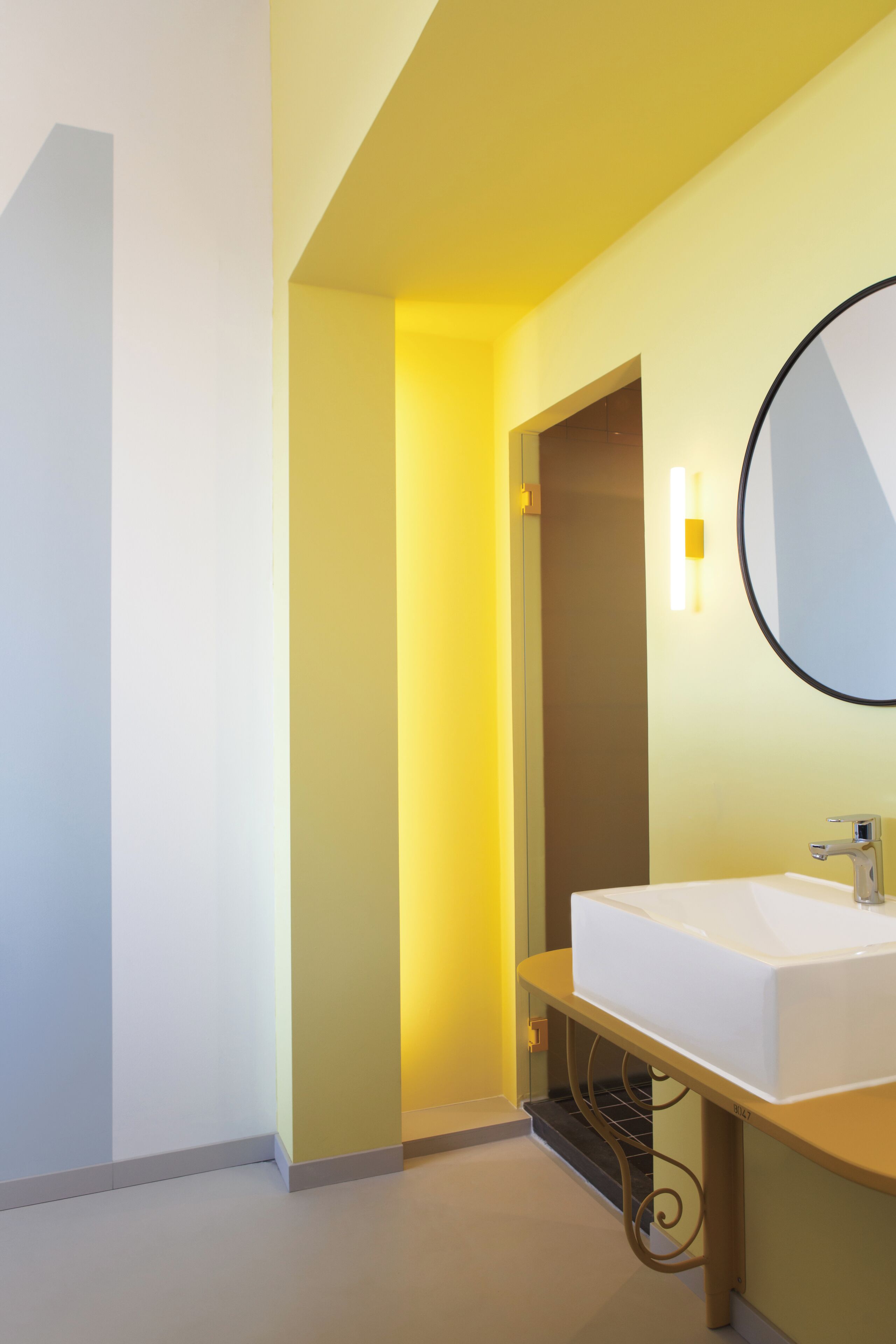 A modern bathroom with a white sink, a round mirror, and a yellow accent wall.