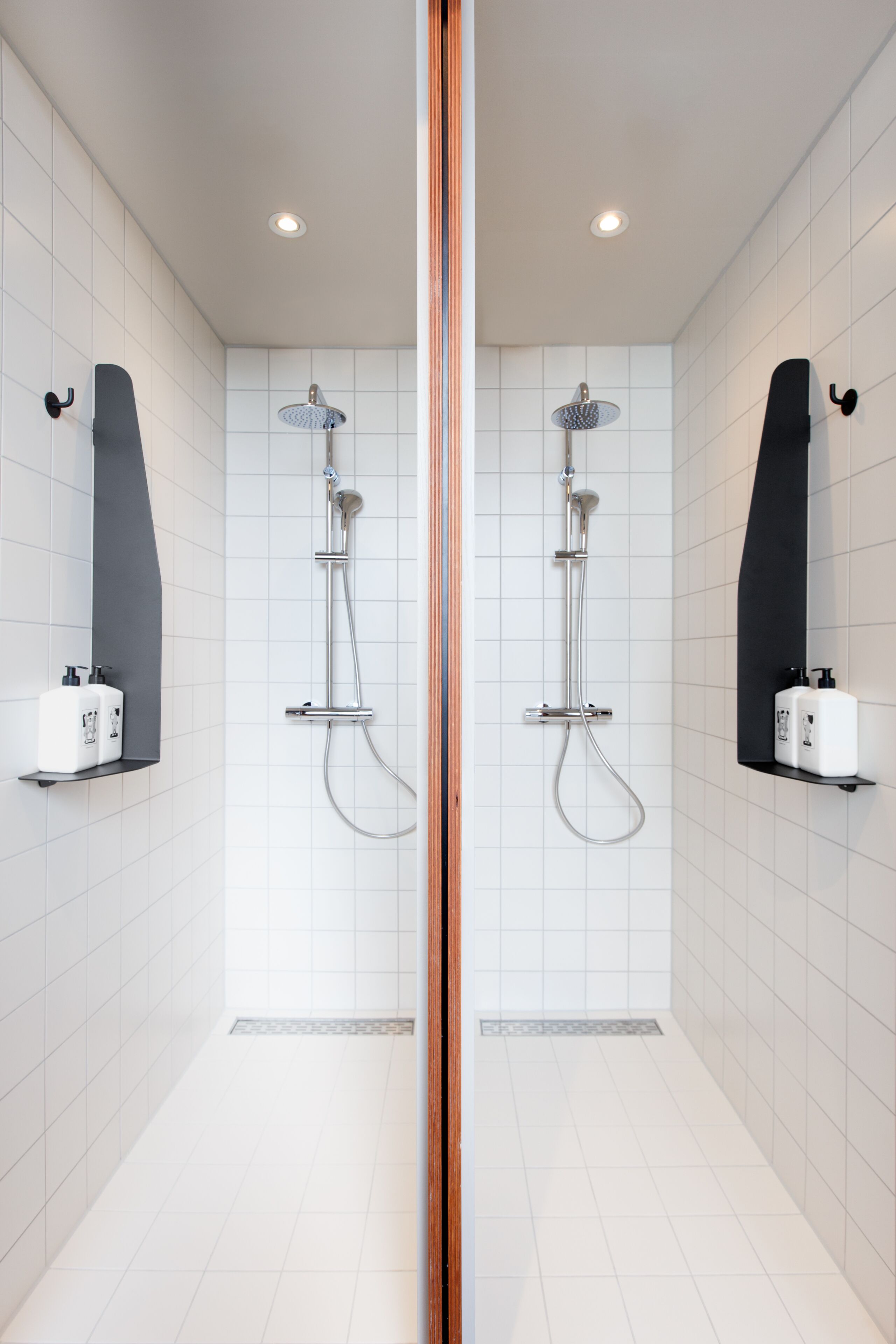 Un baño moderno con paredes de azulejos blancos, dos duchas con estantes negros empotrados y un espejo central.