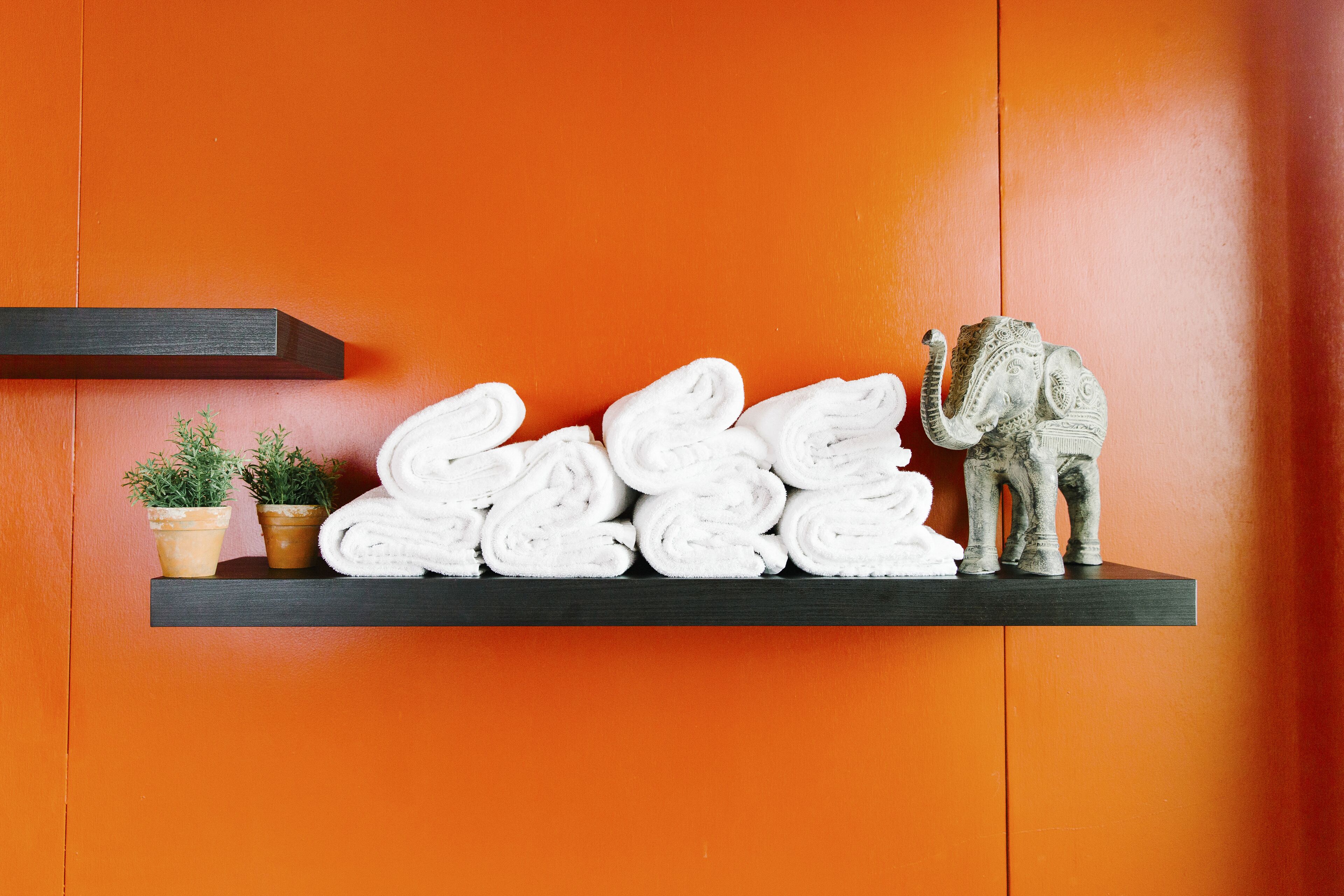 Une étagère avec des serviettes enroulées, deux plantes en pot et une statuette d’éléphant décorative, le tout placé devant un mur orange.