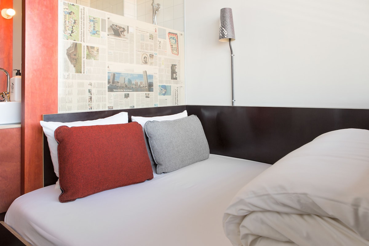 Una cama bien tendida con almohadas blancas y grises, una almohada roja y una colcha blanca. A la derecha, una lámpara de pared y un póster con varias imágenes en la pared detrás de la cama.