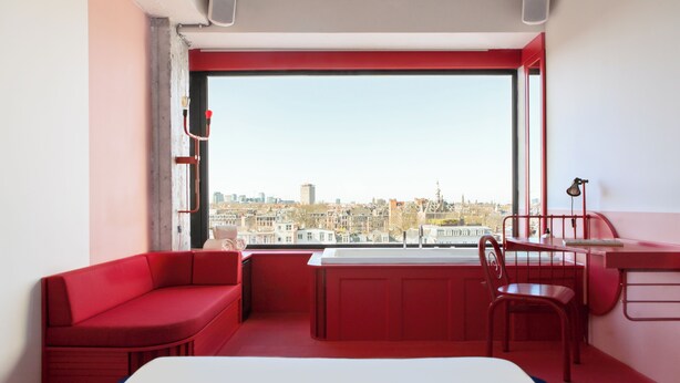 A red-themed room with a city view, a bed, a desk, and a chair.
