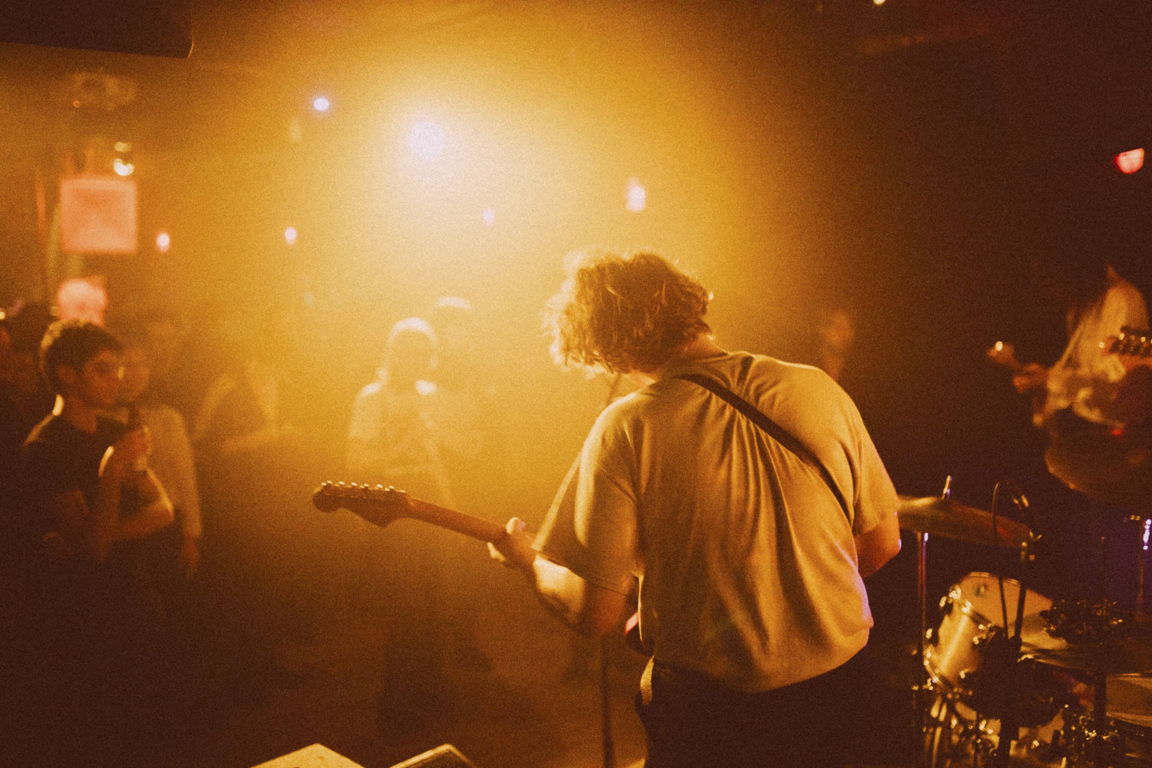 A person playing a guitar on a stage with a drum set in the background.