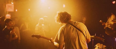 A person playing a guitar on a stage with a drum set in the background.