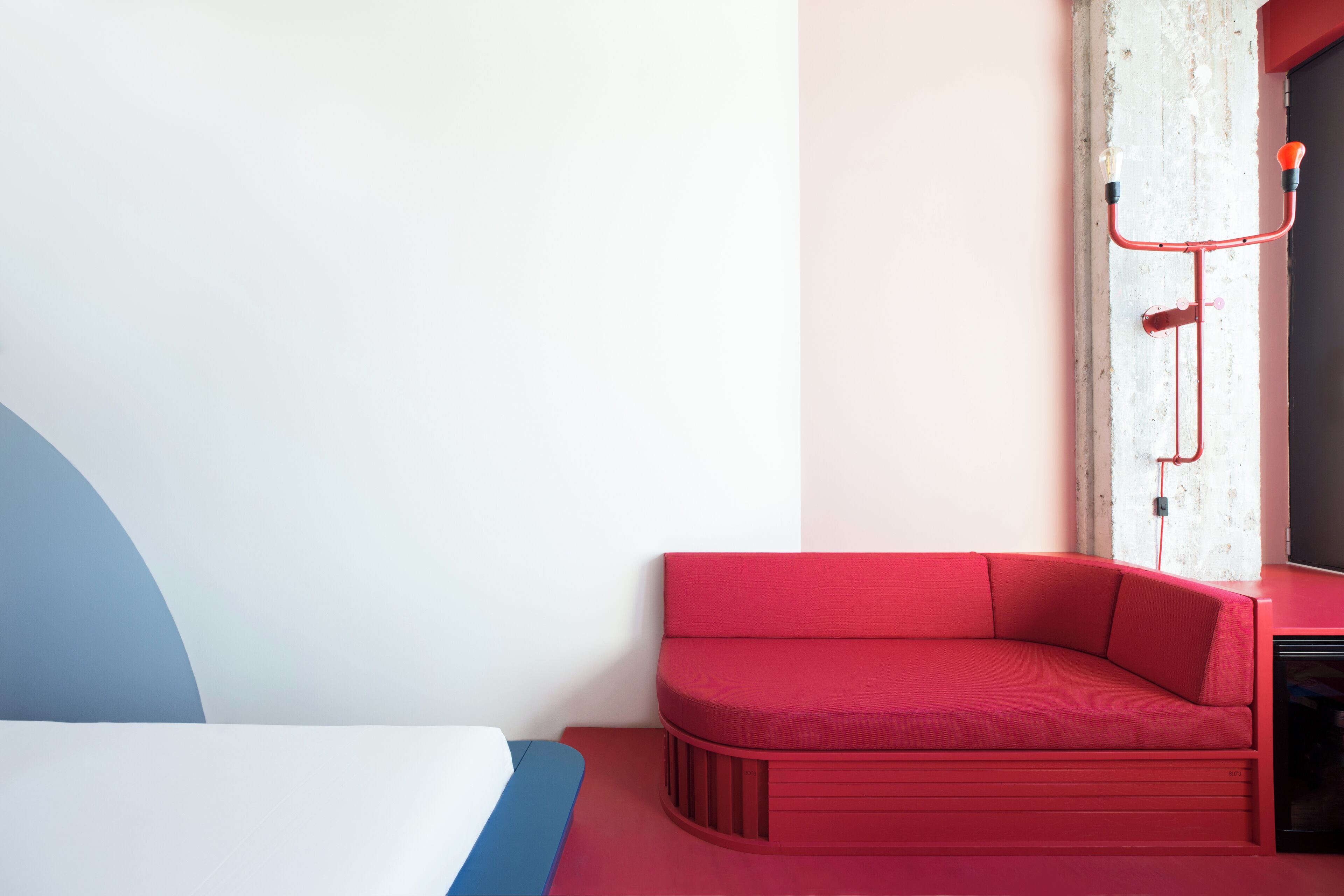 Habitació moderna amb un sofà vermell, un llit amb capçalera blava i blanca, i una paret blanca amb una canonada vermella.
