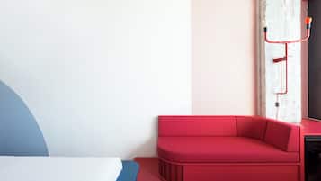 Habitació moderna amb un sofà vermell, un llit amb capçalera blava i blanca, i una paret blanca amb una canonada vermella.