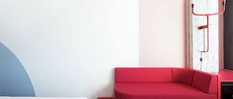 A modern room with a red sofa, a bed with a blue and white headboard, and a white wall with a red pipe.