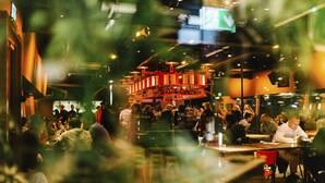 A busy restaurant with multiple dining tables and chairs, a bar area, and a staircase leading to an upper level.