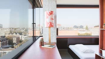 A modern hotel room with a large window, a bed, a desk, and a city view.