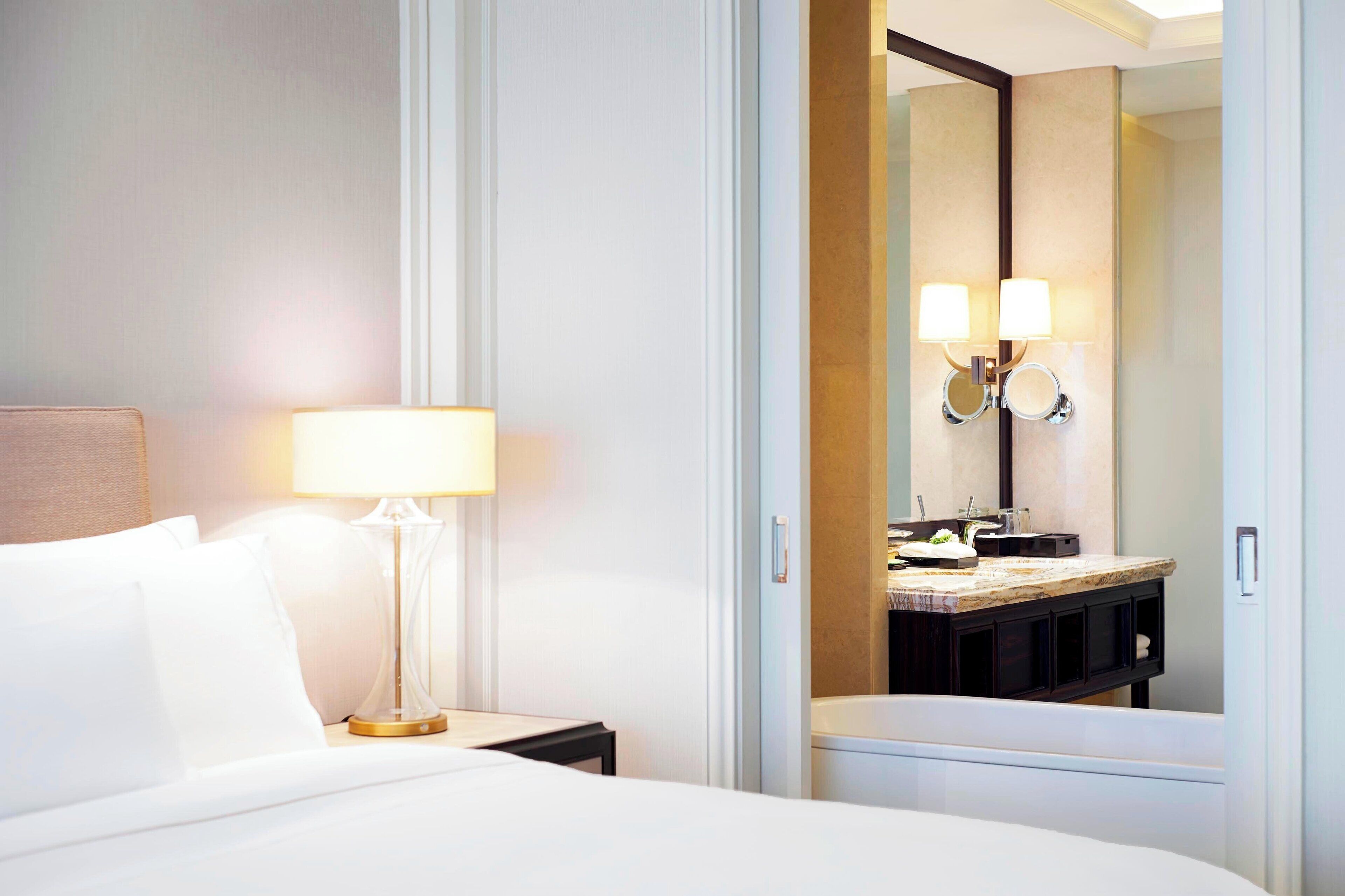 Een hotelkamer met een bed, een nachtkastje met een lamp, een badkamer met een spiegel en een wastafel met aanrecht.