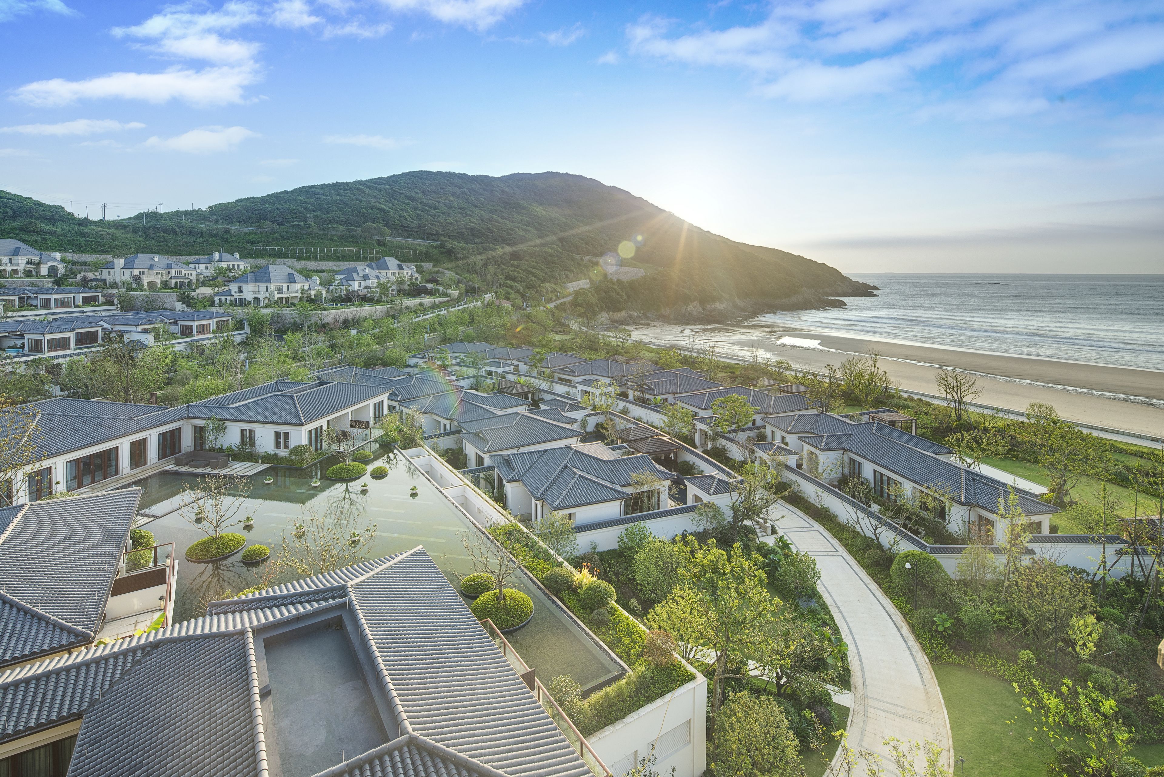 A coastal residential area with modern houses, a beach, and a hilly backdrop.