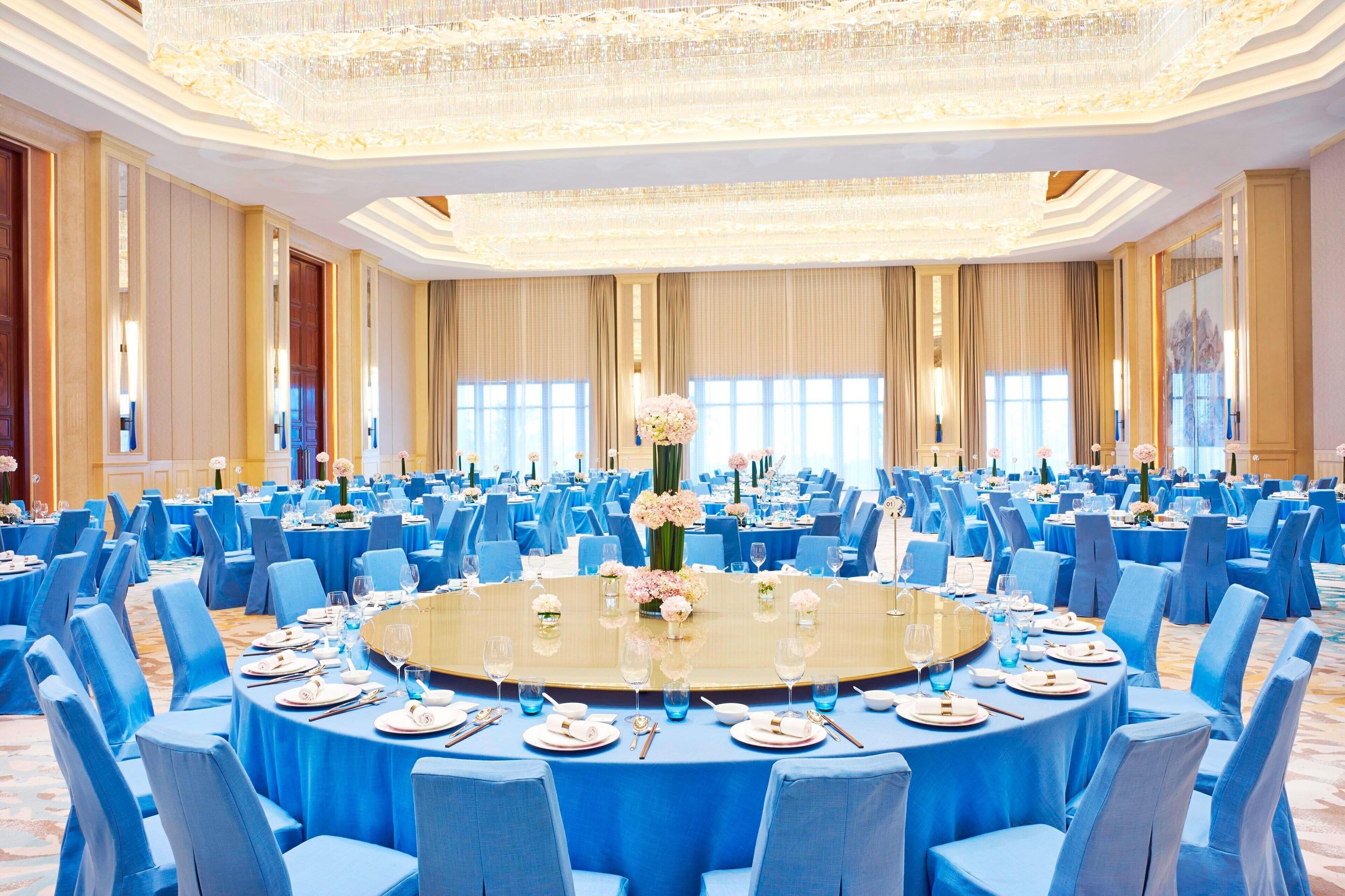 A large conference room with round tables, blue chairs, and a central floral arrangement.