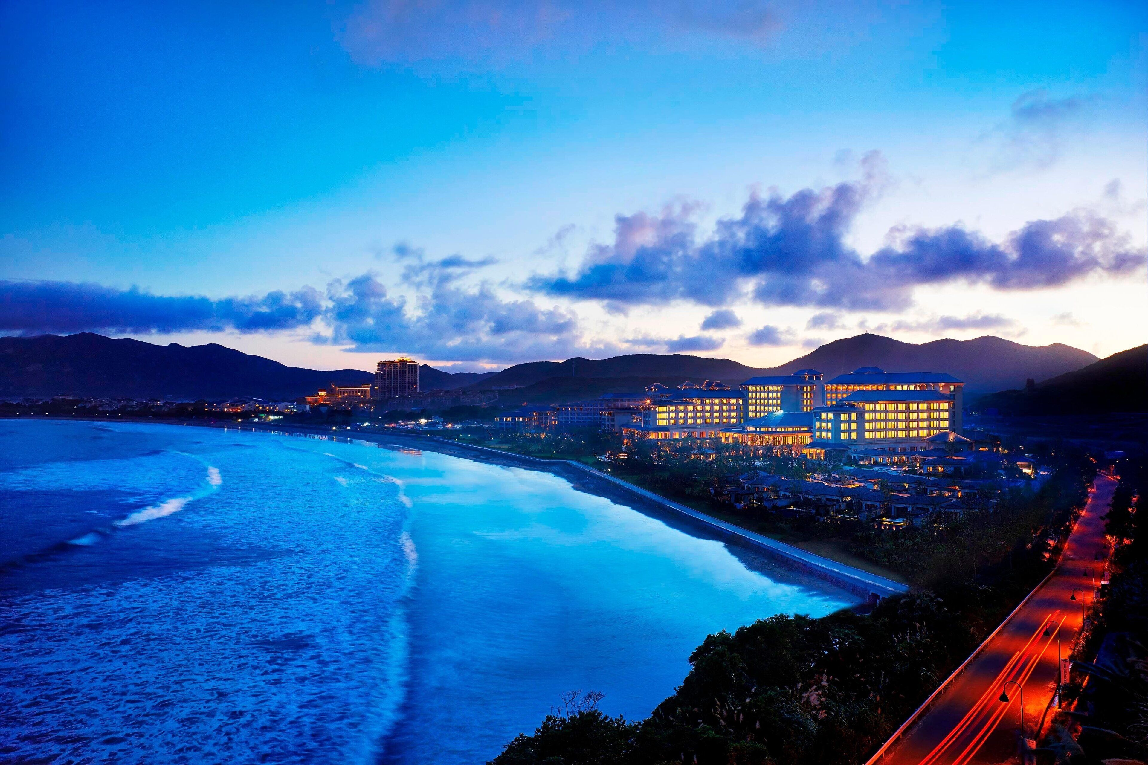 Un hotel costero con vista a la playa, una carretera con luces en movimiento y montañas al fondo.