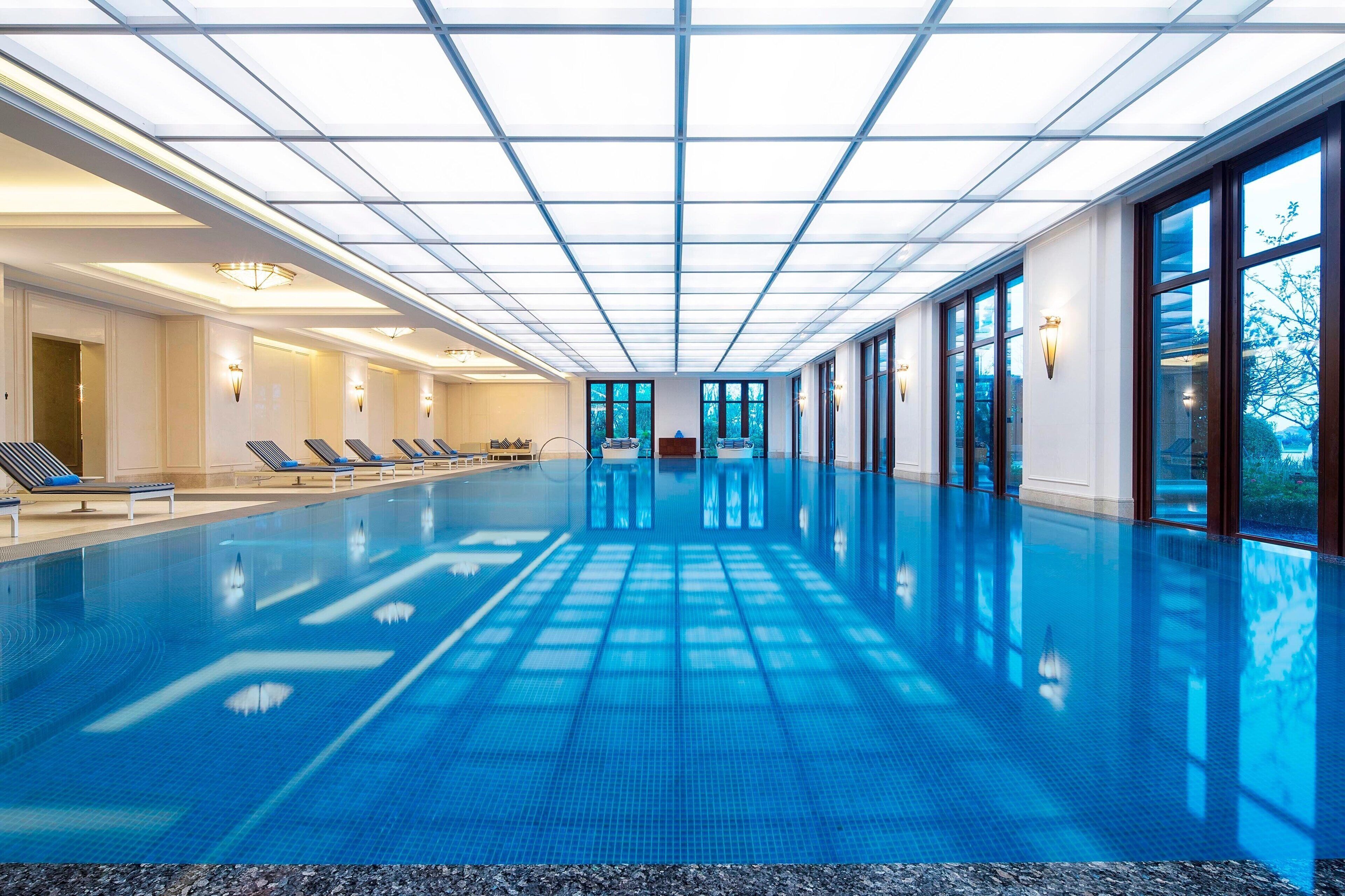 A large indoor swimming pool with clear blue water, sunlit ceiling, and lounge chairs along the sides.