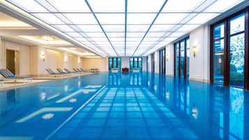 A large indoor swimming pool with clear blue water, sunlit ceiling, and lounge chairs along the sides.
