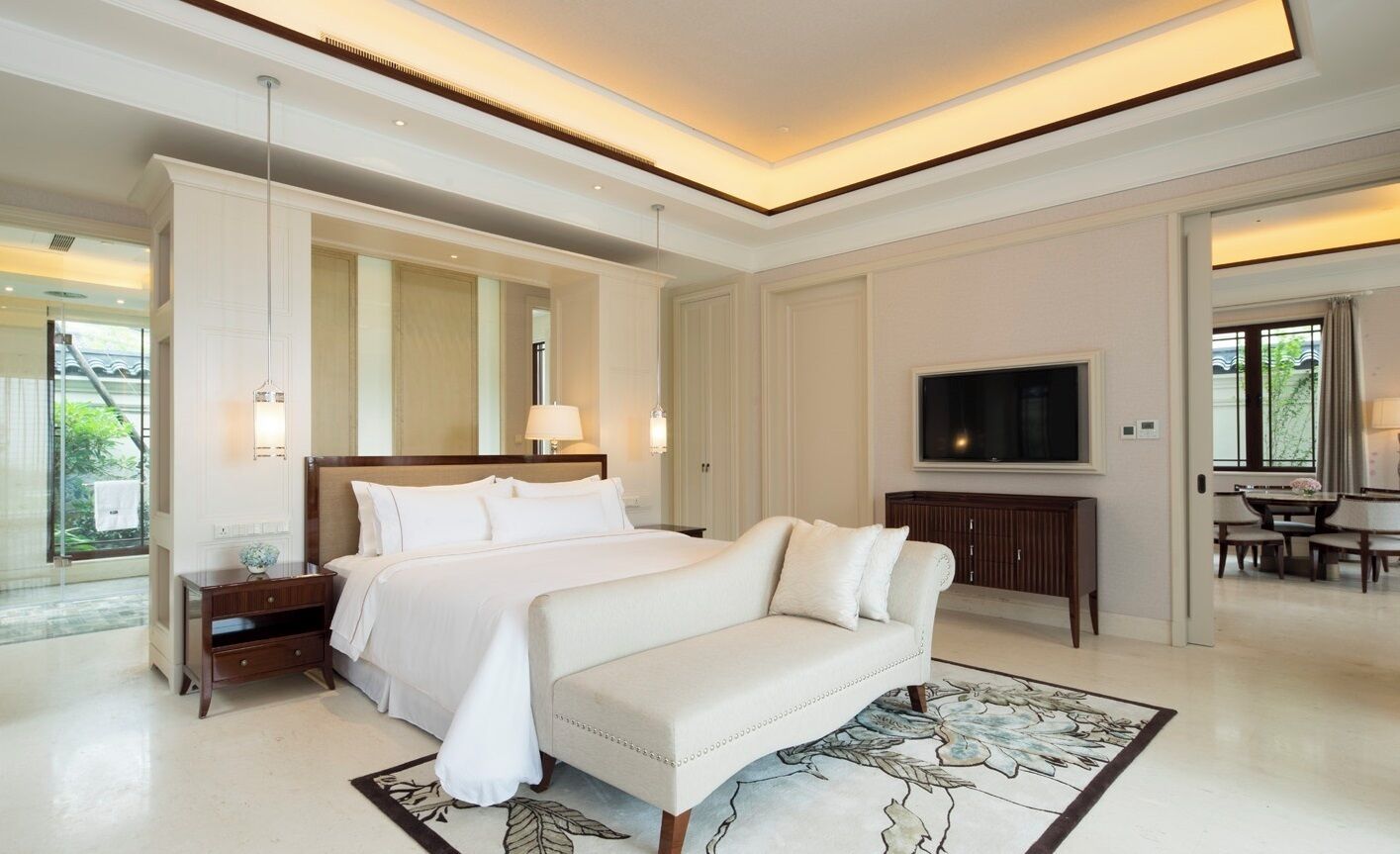 Un dormitorio amplio con una cama grande, un sofá, un televisor y un comedor visible a través de una puerta abierta.