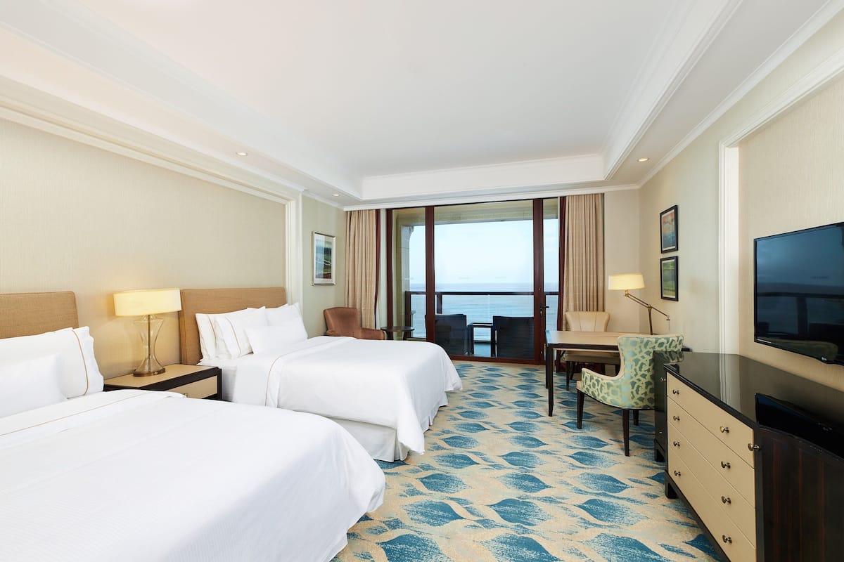Une chambre d’hôtel avec deux lits, un bureau, une chaise, une télévision et une grande fenêtre donnant sur la mer.