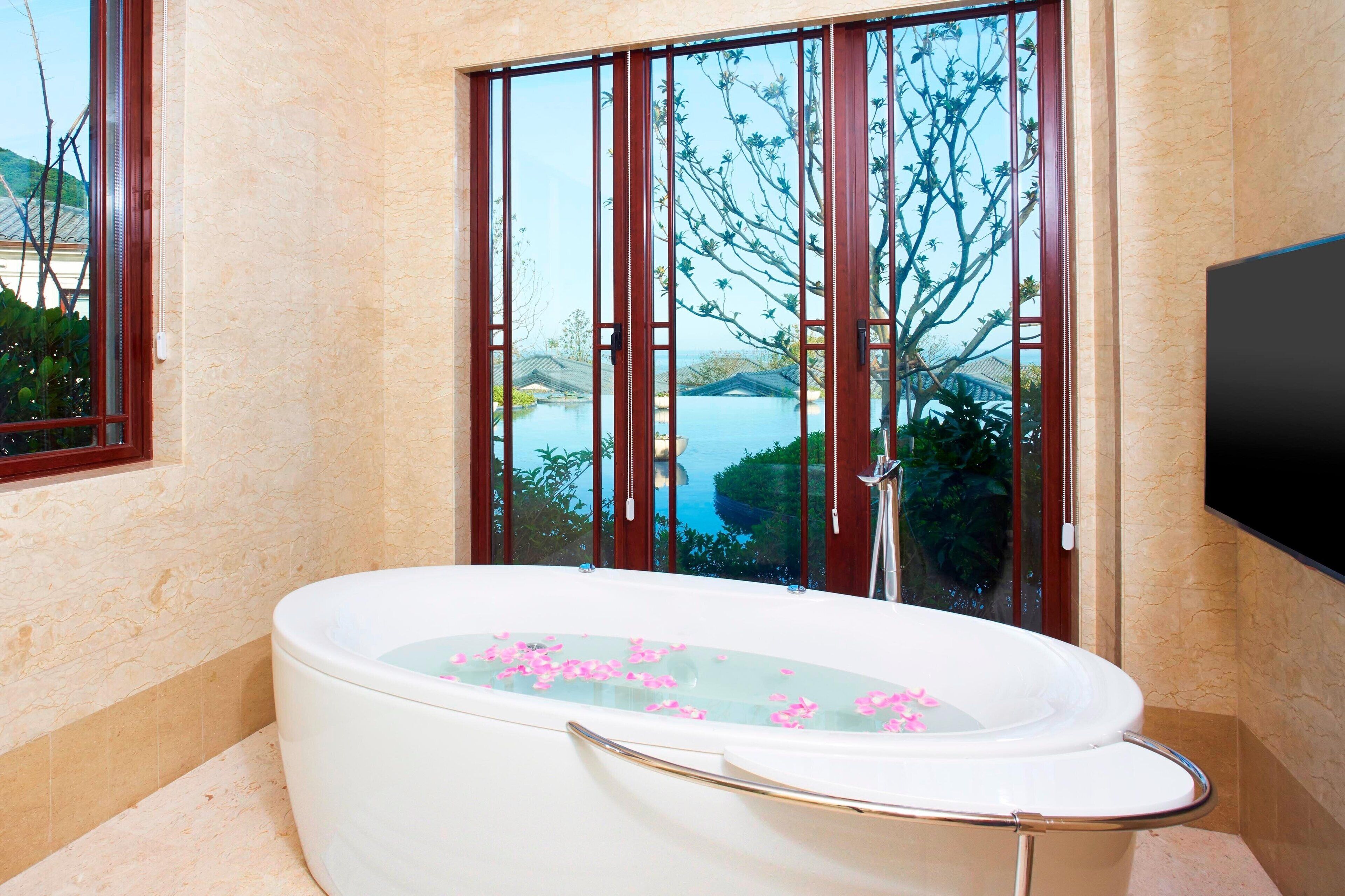 A bathroom with a large bathtub filled with water and rose petals, a window with a view of a body of water and trees, and a flat-screen TV mounted on the wall.