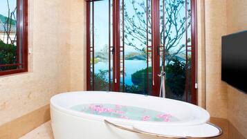 A bathroom with a large bathtub filled with water and rose petals, a window with a view of a body of water and trees, and a flat-screen TV mounted on the wall.