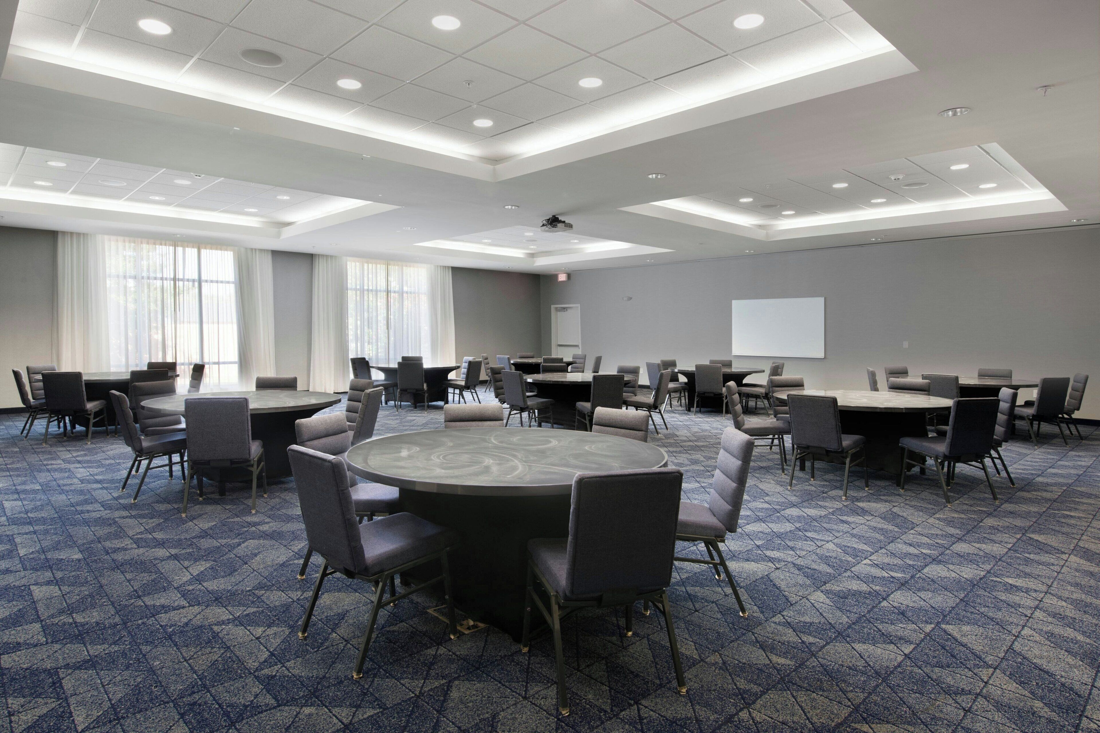 A conference room with multiple tables and chairs, large windows, and a projector screen.