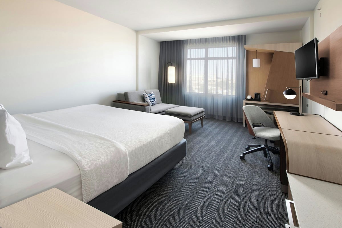 Habitación de hotel con cama, escritorio, silla, televisor y ventana con cortinas.