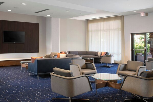Um saguão de hotel com um sofá modular, poltronas, uma TV de tela plana e uma grande janela com cortinas.