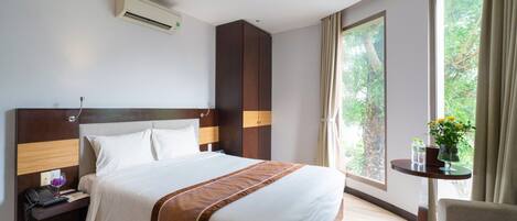 A hotel room with a large bed, a bedside table with a phone and glass, a small table with a vase of flowers, and a window with a view of trees.