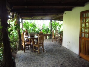 A covered patio area with a stone floor, wooden furniture, and a laptop on a table.