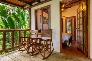 A wooden rocking chair and a bench on a wooden deck with a view of lush greenery.