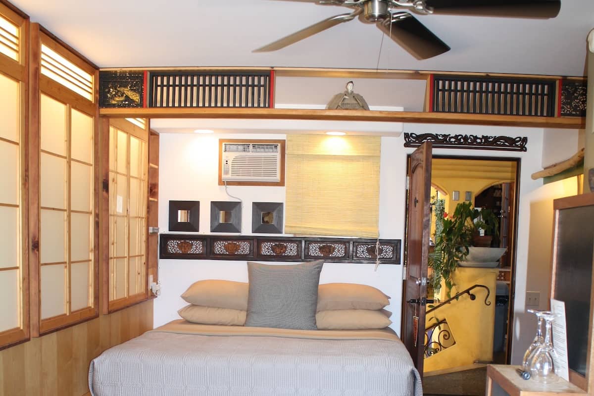 A traditional Japanese-style bedroom with a wooden floor, a bed with a headboard, a ceiling fan, and a sliding door leading to another room.
