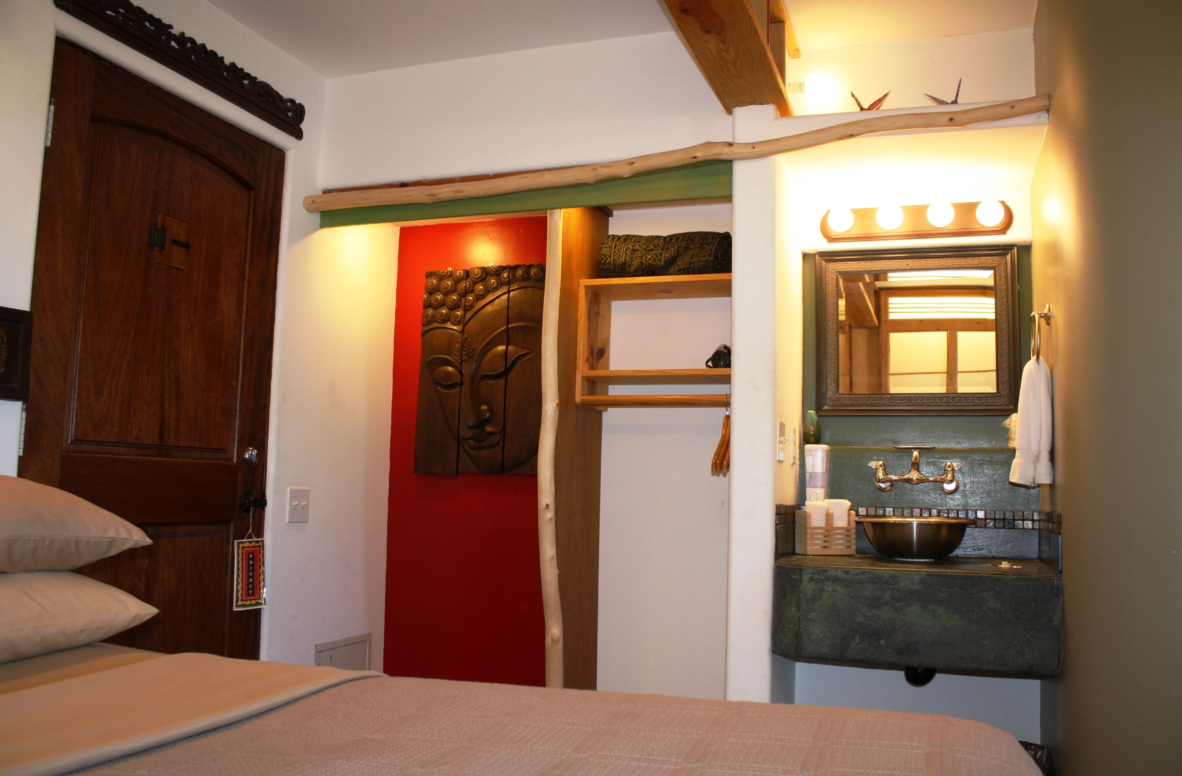 Een slaapkamer met een houten deur, een rode accentmuur, een bed met een beige deken, een houten plankje met toiletartikelen, en een badkamer met een spiegel, wastafel en handdoekrek.