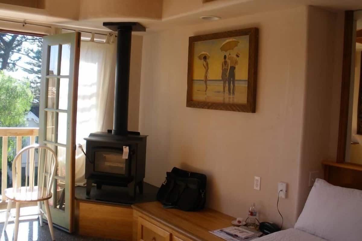 A room with a wood-burning stove, a painting of two people with umbrellas, a chair, a bed, and a wooden bench.