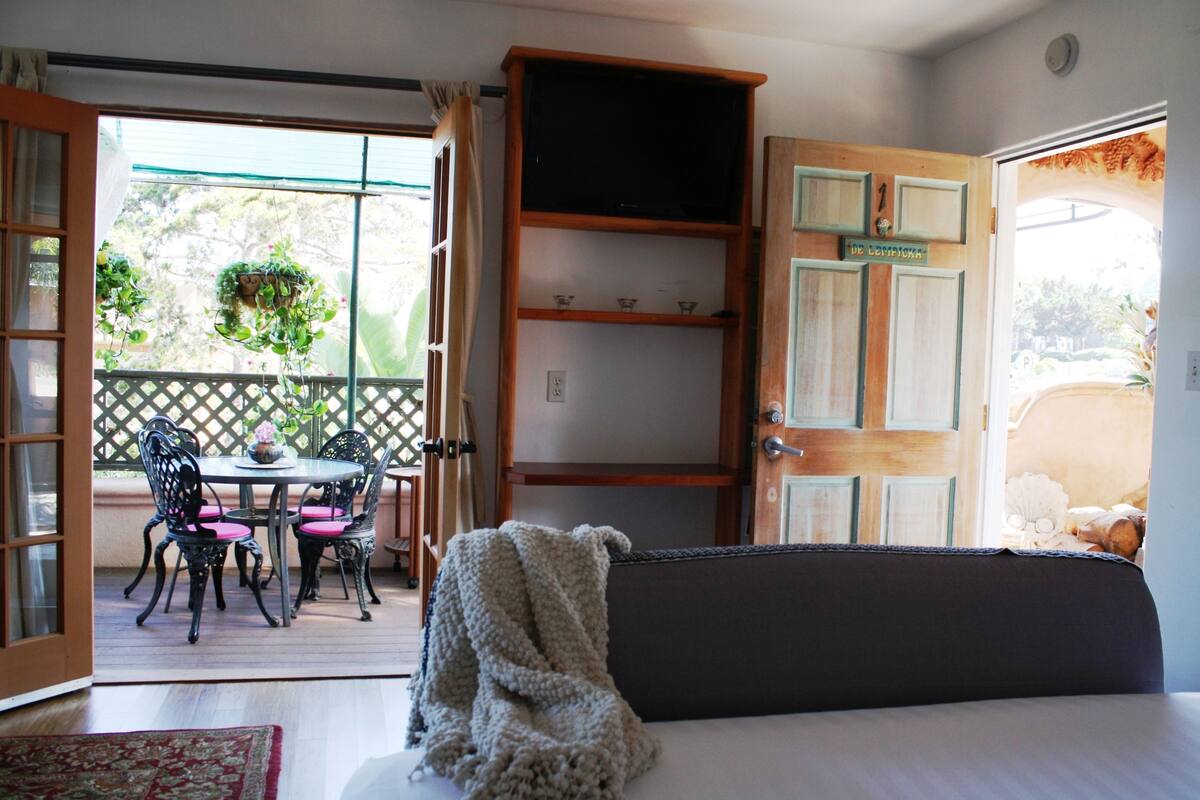 A room with a bed, a window with a view of a garden, a table with chairs, and a door with a sign.