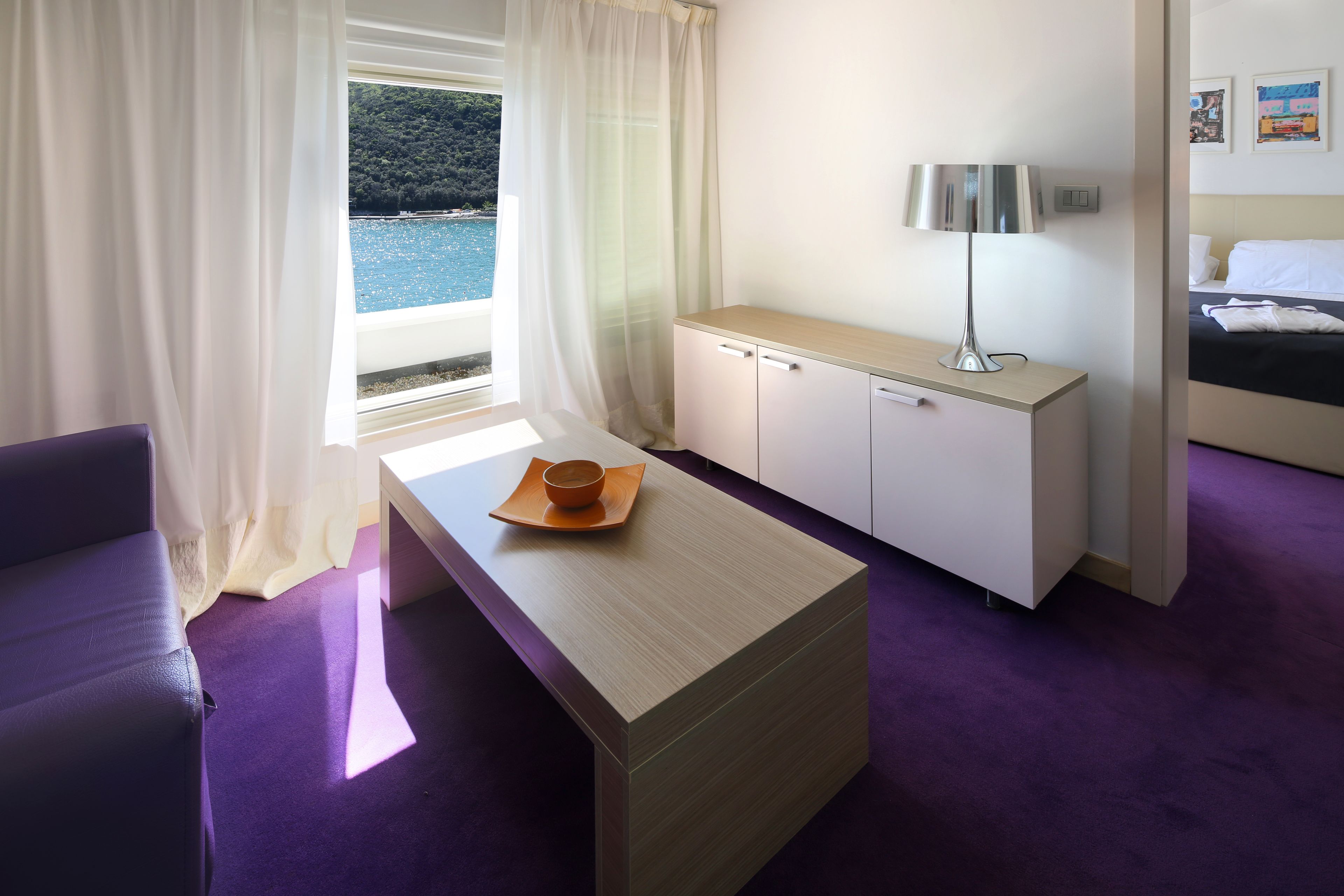 A hotel room with a bed, a desk, a chair, a window with curtains, and a view of the sea.