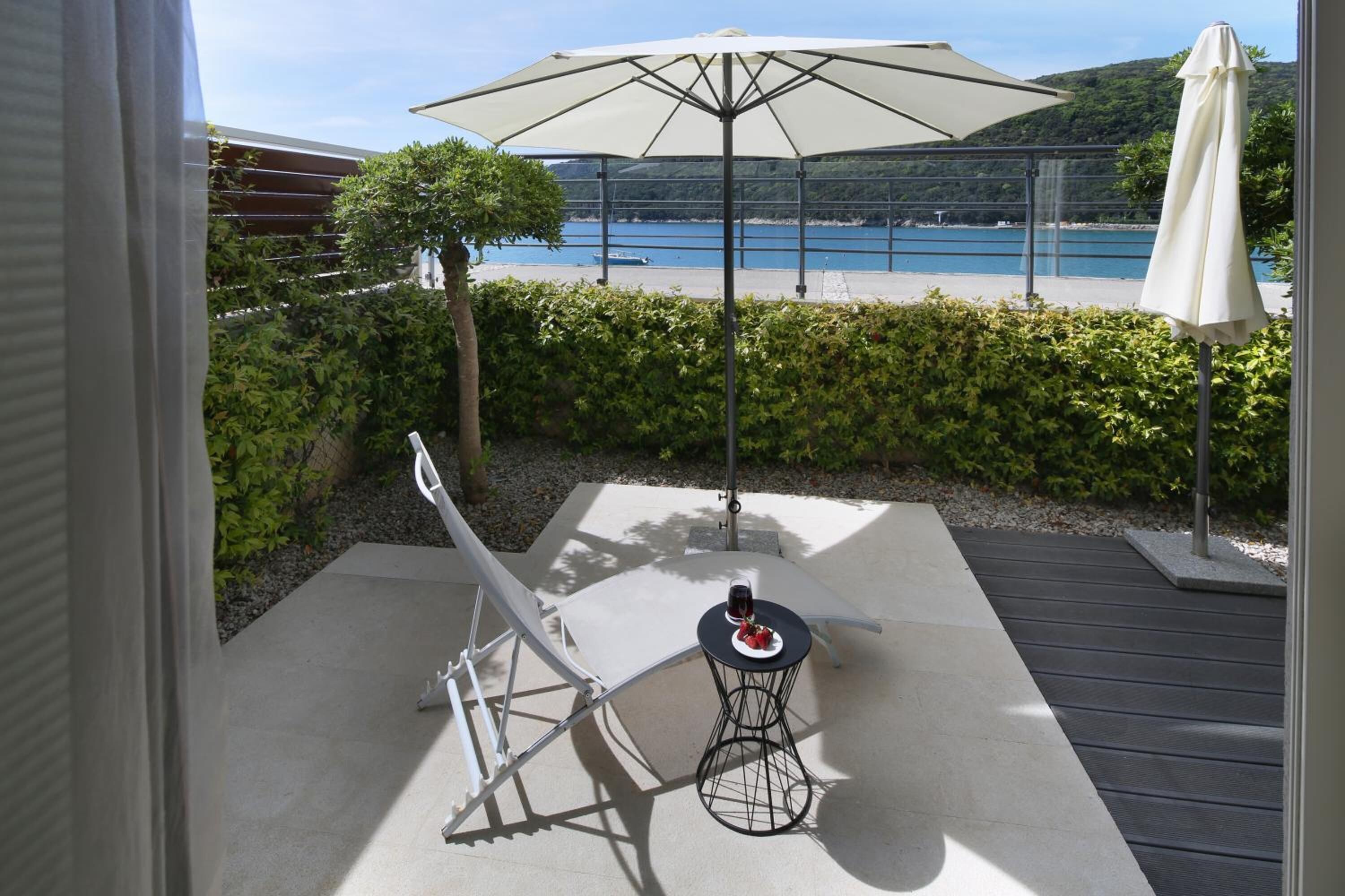 A patio with a lounge chair, a small round table with a drink, and a large umbrella providing shade. A scenic view of a lake and hills is visible in the background.