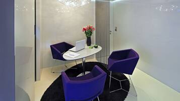 A modern room with a glass partition, a round table with a laptop, two purple chairs, a vase with flowers, and a black rug.