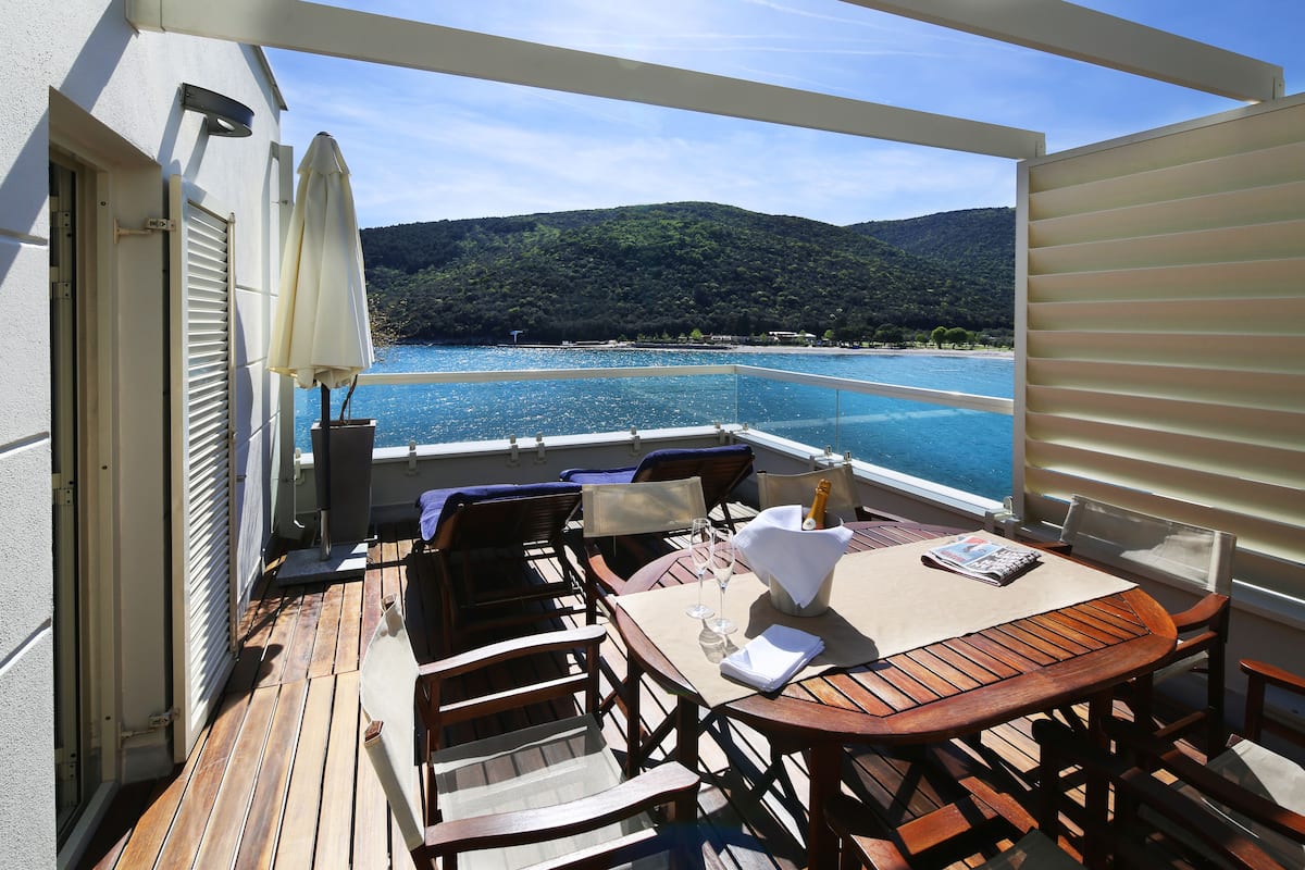 A wooden deck with a table and chairs, overlooking a clear blue sea and a hilly landscape.