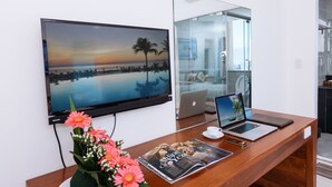 A modern hotel room with a large flat-screen TV mounted on the wall, a wooden desk with a laptop, a magazine, and a vase of flowers. - SOCO Hotel (Da Nang)