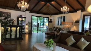 Amplia sala de estar con techo alto, candelabros, un sofá, una mesa con mantel de encaje y vistas a un jardín a través de grandes ventanales.