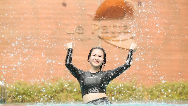 Una persona con traje de neopreno negro, con la palabra "USRONNIE" escrita en él, de pie en el agua con los brazos levantados, y gotas de agua visibles en el aire.