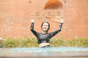 A person in a black wetsuit with the word "USRONNIE" on it, standing in water with arms raised, with water droplets visible in the air.