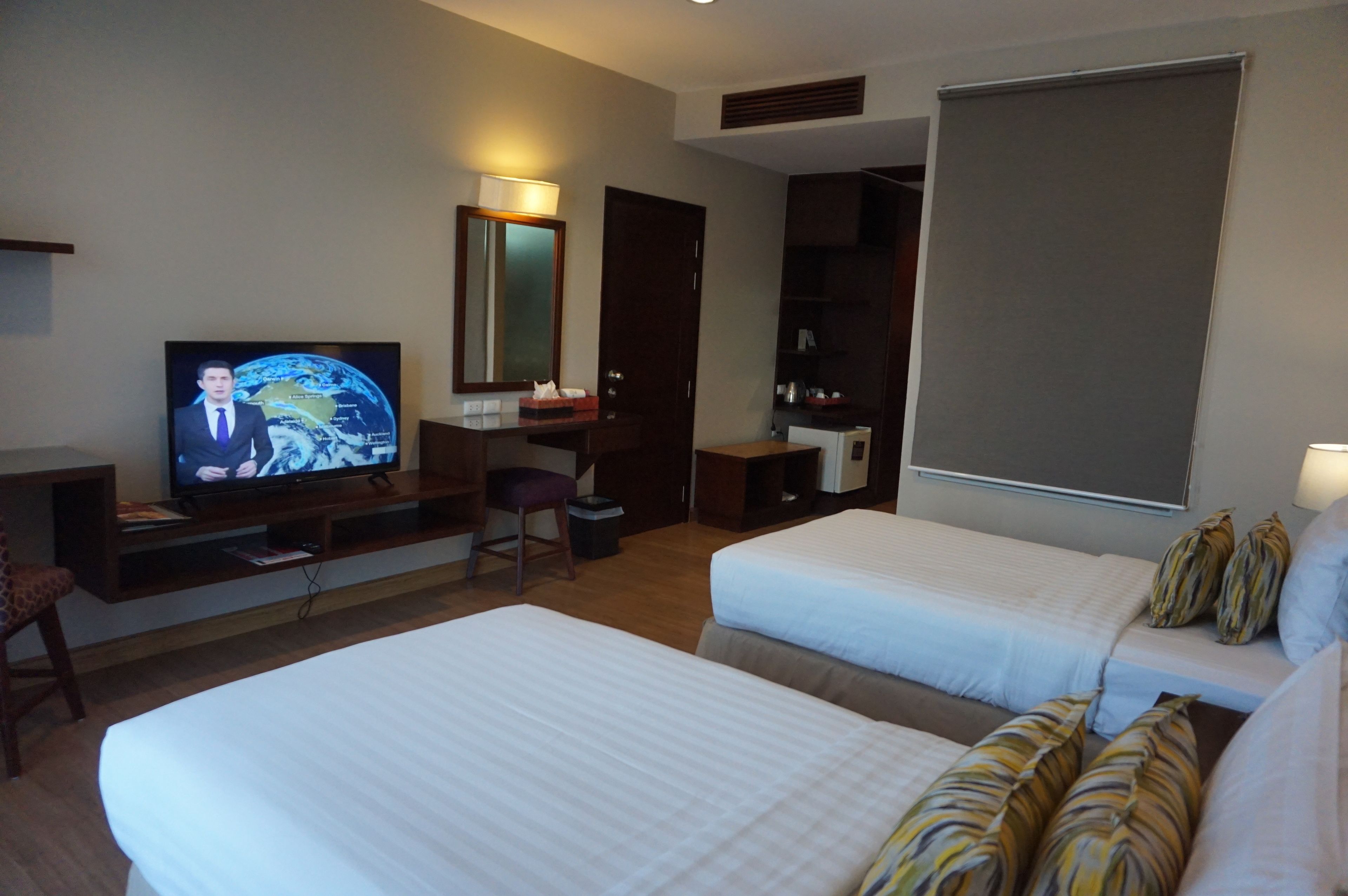 A hotel room with two beds, a desk with a television displaying a news channel, a mirror, a wardrobe, and a large window.