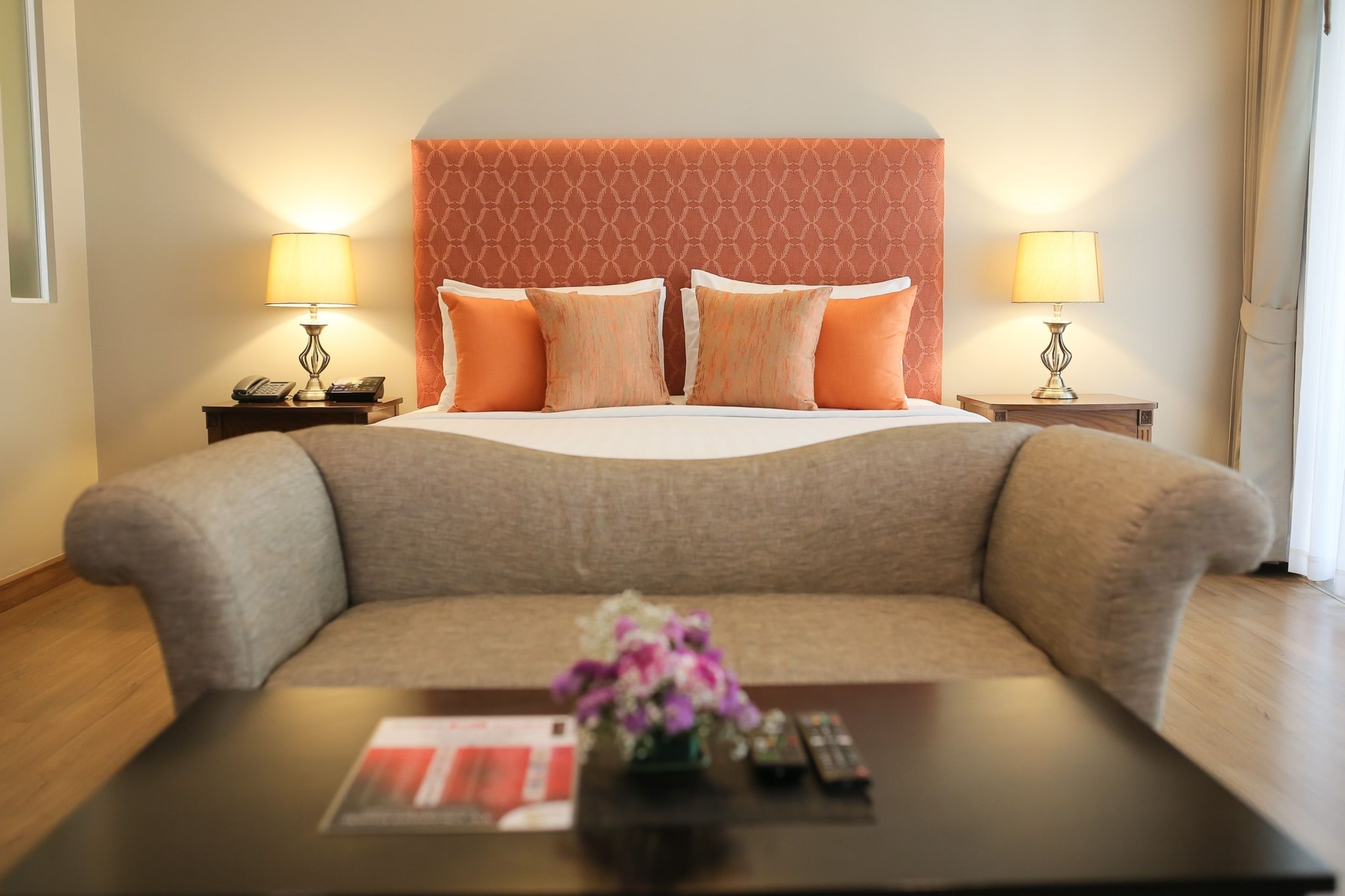 A hotel room with a large bed, two bedside tables with lamps, a sofa, and a coffee table with a remote control and a flower vase.