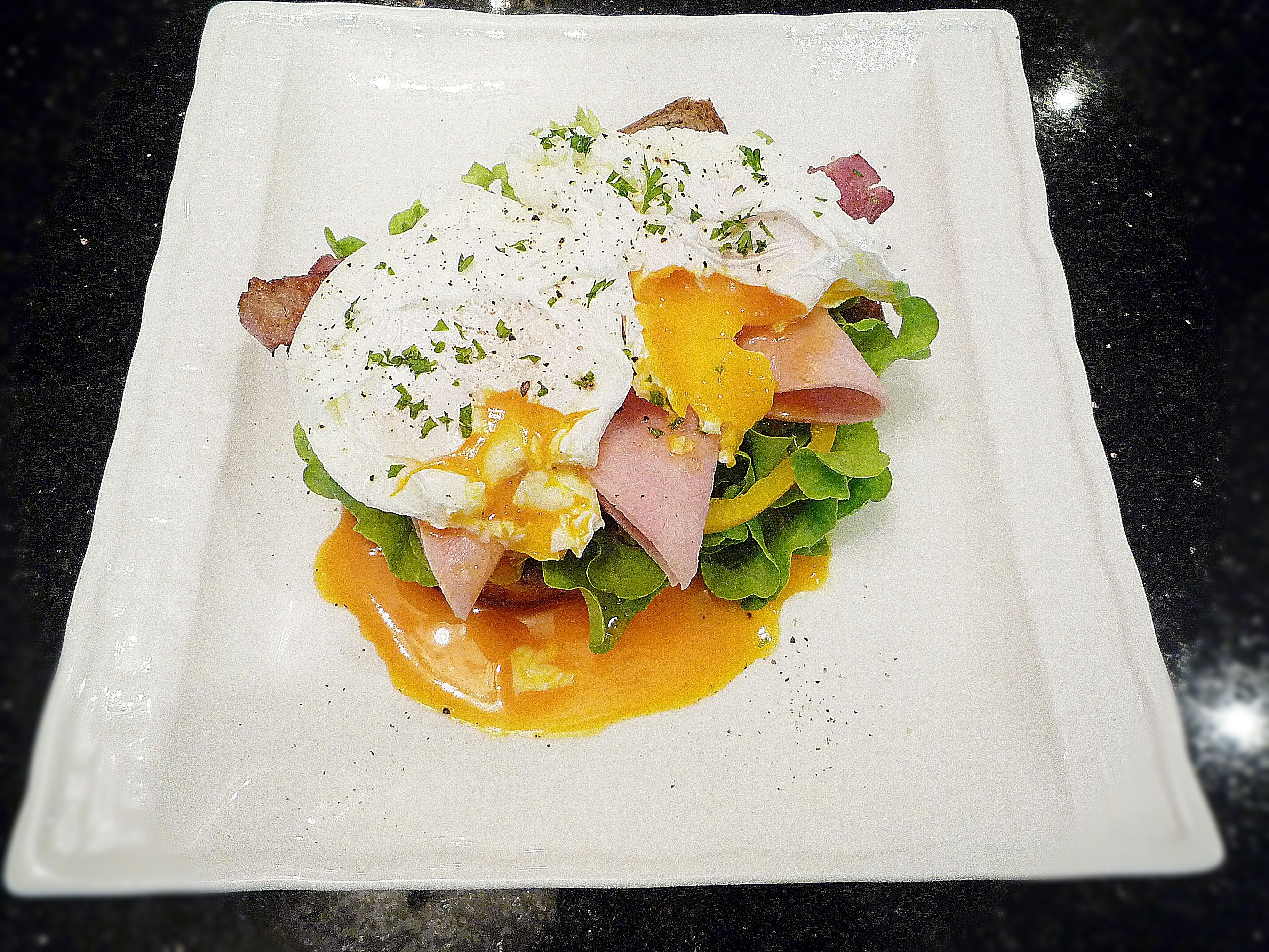 Plato blanco cuadrado con un desayuno que incluye huevos pochados, jamón, queso y verduras, sobre una encimera negra.