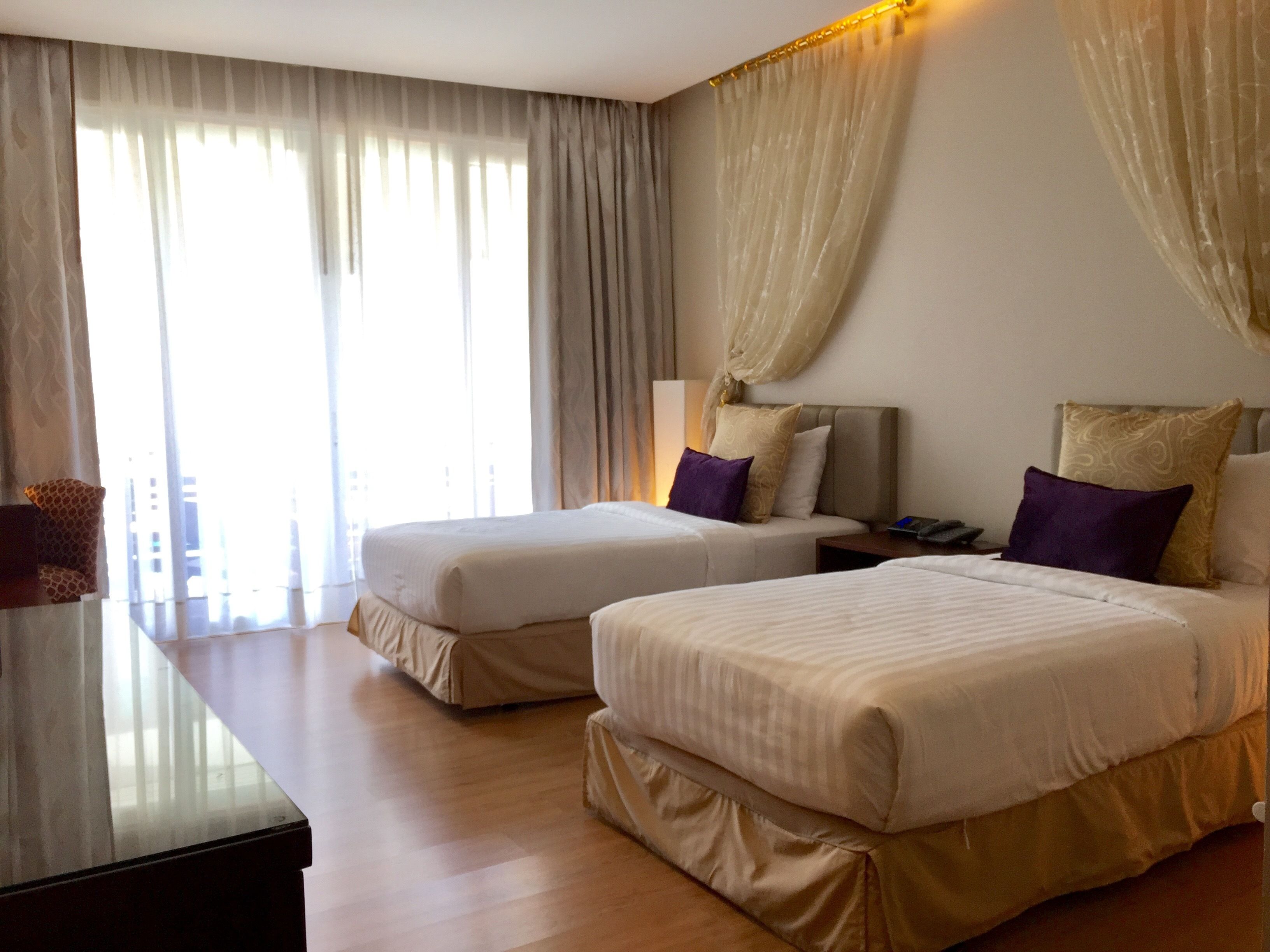 A hotel room with two beds, a nightstand, a lamp, and a large window with curtains.