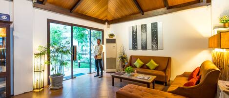 A spacious living area with a wooden ceiling, a brown sofa, a coffee table, and a large window offering a view of greenery outside.