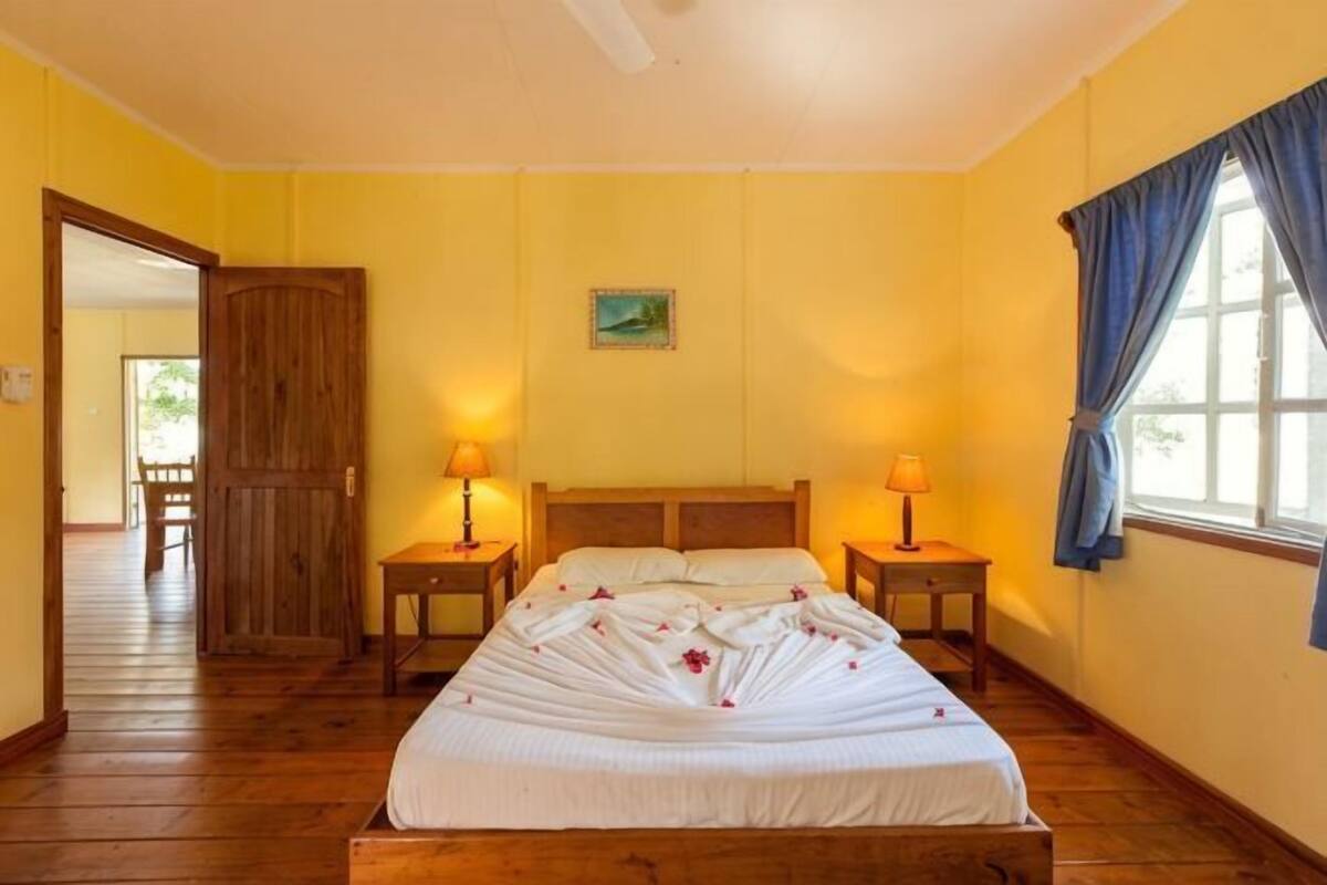 A bedroom with a wooden bed, two bedside tables, a window with curtains, and a door leading to another room.