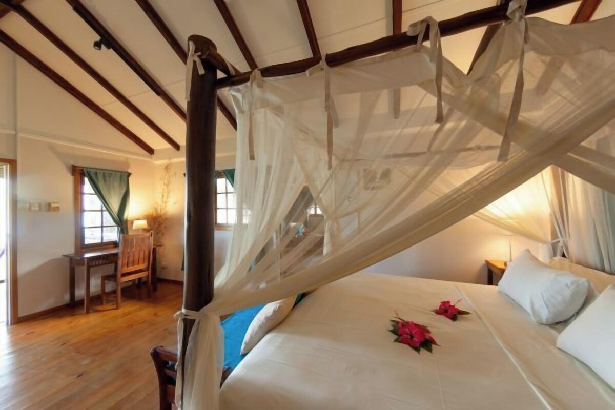 A bedroom with a four-poster bed, wooden beams on the ceiling, and a desk with a chair in the corner.