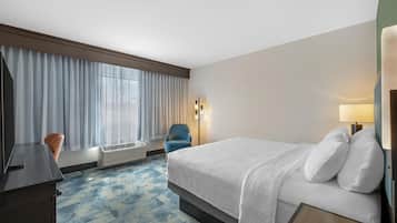 Una habitación de hotel con cama, escritorio, silla, lámpara y ventana con cortinas.